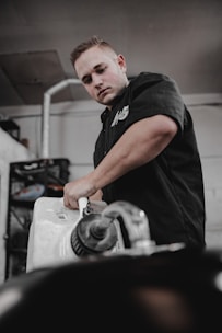 A person in a black shirt is working with a large plastic container, possibly a gasoline or liquid container, inside a workshop or garage. The setting includes industrial elements like metal pipes and shelves in the background.