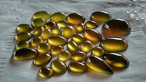 A collection of polished, translucent stones or beads with a glossy surface, arranged on a textured white surface. The stones display a rich, golden-amber hue with subtle gradients, creating a smooth and reflective appearance.
