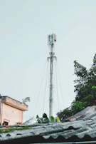 A telecommunications tower rises prominently against a pale sky. It is surrounded by lush green foliage and the edge of a building is visible on the left side. The scene also includes parts of a rooftop at the bottom.