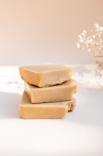 Three stacked bars of handmade soap with a natural, beige color sit on a soft white surface. A delicate sprig of dried flowers is visible in the background, adding an elegant touch to the minimalistic composition.