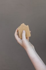 A hand holding a piece of brown soap against a plain background. The soap has a natural, handmade appearance with a slightly uneven surface.