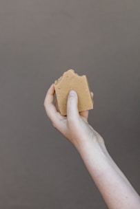 A hand holding a piece of brown soap against a plain background. The soap has a natural, handmade appearance with a slightly uneven surface.