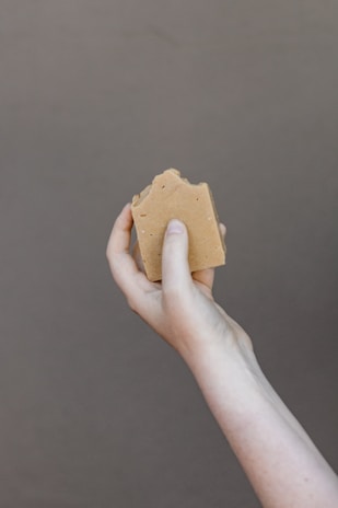 A hand holding a piece of brown soap against a plain background. The soap has a natural, handmade appearance with a slightly uneven surface.