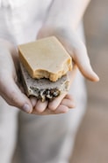 A pair of hands gently holding two pieces of artisan soap. One soap is a plain, beige color, while the other is speckled with natural inclusions, possibly herbs or seeds, and features some decorative elements. The lighting is soft and natural, focusing on the soaps’ textures.