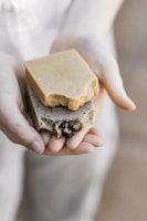A pair of hands gently holding two pieces of artisan soap. One soap is a plain, beige color, while the other is speckled with natural inclusions, possibly herbs or seeds, and features some decorative elements. The lighting is soft and natural, focusing on the soaps’ textures.