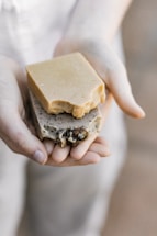 A pair of hands gently holding two pieces of artisan soap. One soap is a plain, beige color, while the other is speckled with natural inclusions, possibly herbs or seeds, and features some decorative elements. The lighting is soft and natural, focusing on the soaps’ textures.