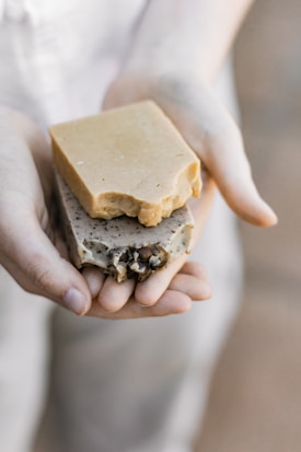 A pair of hands gently holding two pieces of artisan soap. One soap is a plain, beige color, while the other is speckled with natural inclusions, possibly herbs or seeds, and features some decorative elements. The lighting is soft and natural, focusing on the soaps’ textures.