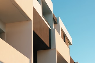 A modern architectural building with sharp lines and contrasting colors. The structure features large rectangular facades with a smooth finish, highlighted by natural lighting. The clear blue sky serves as a backdrop, enhancing the building's geometric design.