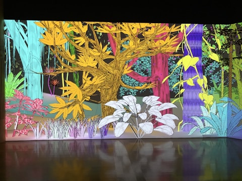 A vibrant and colorful illustration of a forest scene with various stylized trees and plants. The artwork features a yellow tree with elaborate branches in the center, surrounded by other trees in shades of blue, pink, and green. Abstract vegetation with detailed leaves and textures fills the foreground and background.