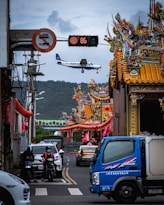 An airplane is flying low over a street with vibrant, traditional Asian temple architecture featuring intricate designs and colorful details. Below, a busy street scene includes cars, motorcycles, and a blue truck. A digital traffic sign is visible, displaying the number 86.