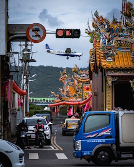 An airplane is flying low over a street with vibrant, traditional Asian temple architecture featuring intricate designs and colorful details. Below, a busy street scene includes cars, motorcycles, and a blue truck. A digital traffic sign is visible, displaying the number 86.