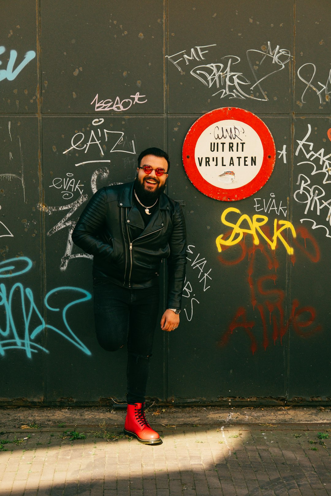man in black leather jacket standing beside wall with red and white no smoking sign