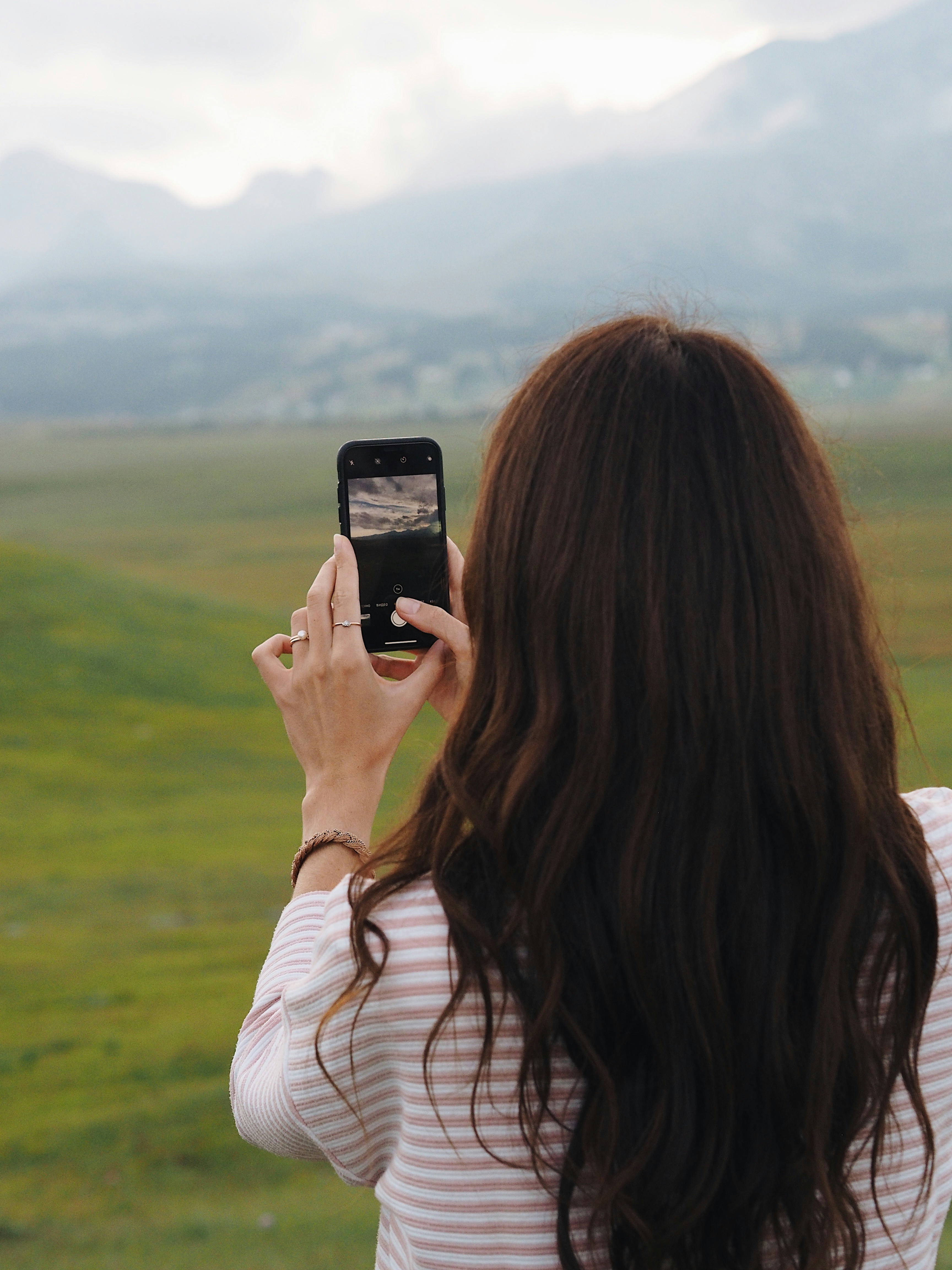 A back view of a girl with long curly hair, who takes pictures of picturesque green mountains and broadcasts on Instagram.