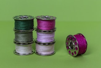 Six spools of thread stacked in groups of three against a bright green background. Each spool is metallic, with threads of various colors including dark green, pink, light green, and white. One spool is placed separately to the right.