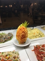 An intricately carved fruit centerpiece resembling a rose, set on a white plate. Surrounding the centerpiece are various dishes, including a pasta salad and a mix of vegetables, displayed on a buffet line. The background is a dimly lit restaurant with people dining.