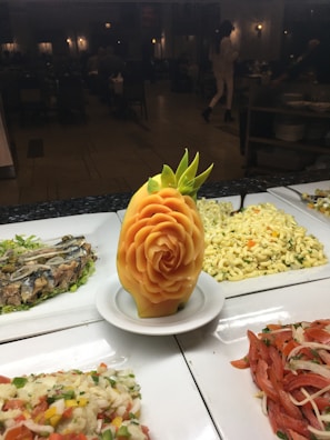 An intricately carved fruit centerpiece resembling a rose, set on a white plate. Surrounding the centerpiece are various dishes, including a pasta salad and a mix of vegetables, displayed on a buffet line. The background is a dimly lit restaurant with people dining.