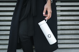 A person wearing dark clothing is holding a sleek, white cylindrical bottle with a strap in their hand. The background consists of horizontal wooden slats, adding a modern, minimalist look to the scene.