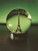 A snow globe featuring a miniature Eiffel Tower inside, set against a dark green backdrop. The base of the globe is labeled with the word 'Paris' in white letters.