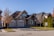 A suburban house with a brick facade and double garage doors. The roof is dark, and there are several bare trees and green bushes surrounding the property. A for sale sign is visible on the front lawn.