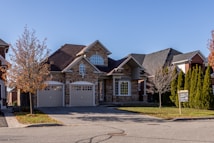 A suburban house with a brick facade and double garage doors. The roof is dark, and there are several bare trees and green bushes surrounding the property. A for sale sign is visible on the front lawn.