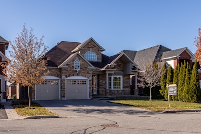 A suburban house with a brick facade and double garage doors. The roof is dark, and there are several bare trees and green bushes surrounding the property. A for sale sign is visible on the front lawn.