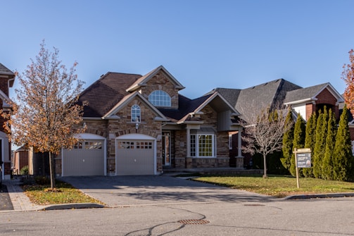 A suburban house with a brick facade and double garage doors. The roof is dark, and there are several bare trees and green bushes surrounding the property. A for sale sign is visible on the front lawn.