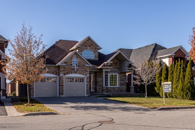 A suburban house with a brick facade and double garage doors. The roof is dark, and there are several bare trees and green bushes surrounding the property. A for sale sign is visible on the front lawn.