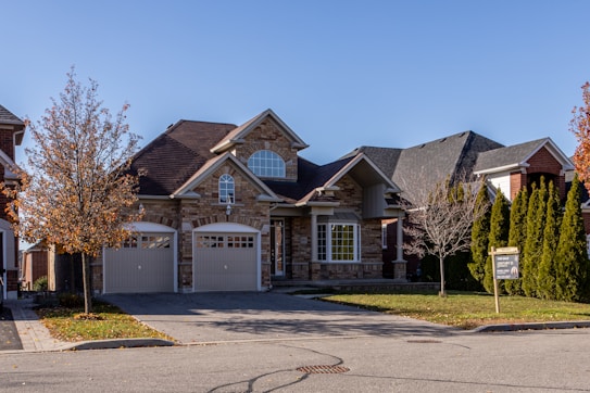 A suburban house with a brick facade and double garage doors. The roof is dark, and there are several bare trees and green bushes surrounding the property. A for sale sign is visible on the front lawn.