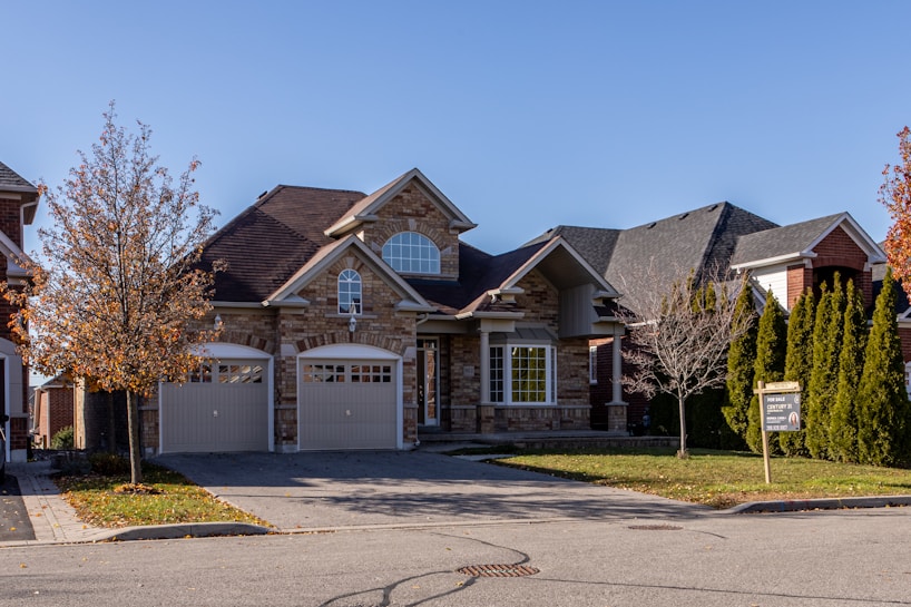 A suburban house with a brick facade and double garage doors. The roof is dark, and there are several bare trees and green bushes surrounding the property. A for sale sign is visible on the front lawn.