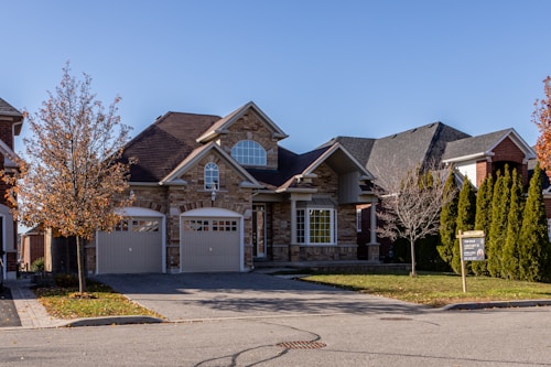 A suburban house with a brick facade and double garage doors. The roof is dark, and there are several bare trees and green bushes surrounding the property. A for sale sign is visible on the front lawn.
