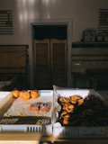A cozy bakery setting with trays of assorted pastries on display. The left tray holds a few pastries including some topped with fruit and powdered sugar, while the right tray is stacked with spiral pastries. Sunlight streams through an unseen window, illuminating the pastries and creating a warm glow.