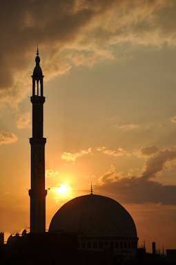 A mosque silhouette against a sunset sky with clouds scattered across the horizon. The sun is partially hidden, casting warm orange and golden hues across the scene. The mosque's dome and a tall minaret are clearly visible.