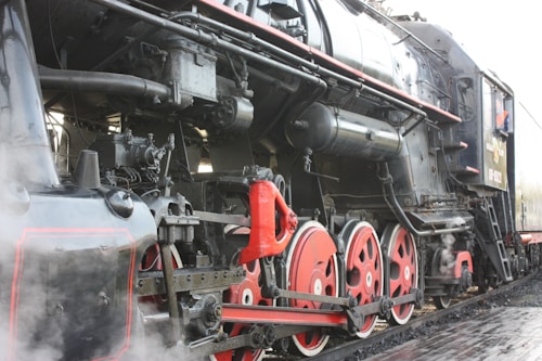 A close-up view of a vintage steam locomotive, showcasing its large red and black wheels, intricate mechanical components, and steam emitting from the engine. The locomotive appears to be stationary on a set of tracks.