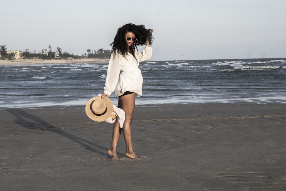 woman in white long sleeve shirt and white skirt holding brown hat standing on beach during