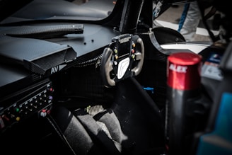 The interior of a race car featuring a steering wheel with multiple buttons and a screen. Surrounding the steering wheel are various controls and displays. The seat appears to be made from a racing material, and a red component labeled 'ALEX' is visible to the right.