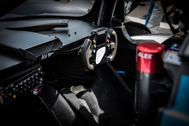 The interior of a race car featuring a steering wheel with multiple buttons and a screen. Surrounding the steering wheel are various controls and displays. The seat appears to be made from a racing material, and a red component labeled 'ALEX' is visible to the right.