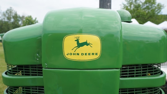 A close-up of the front of a green John Deere tractor displaying the brand logo prominently on the hood, with background elements including grass, trees, and some white structures.