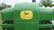 A close-up of the front of a green John Deere tractor displaying the brand logo prominently on the hood, with background elements including grass, trees, and some white structures.