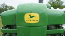 A close-up of the front of a green John Deere tractor displaying the brand logo prominently on the hood, with background elements including grass, trees, and some white structures.