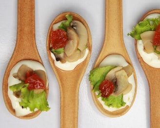Four edible spoons topped with a mixture of ingredients, including slices of mushrooms, a dollop of white creamy sauce, small pieces of red bell pepper, and fresh green lettuce. The spoons are arranged side by side on a white background.