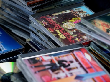 A collection of stacked compact discs (CDs) with colorful album covers, including one prominent with psychedelic art. The CDs are arranged haphazardly, suggesting a music collection. The visible album cover depicts vivid floral and band imagery, hinting at classic rock music.