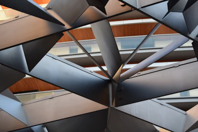 An abstract architectural design featuring a complex pattern of intersecting metallic beams. The structure is layered with a geometric arrangement, highlighting the contrast between the metallic gray and the warm wooden and glass paneling in the background.