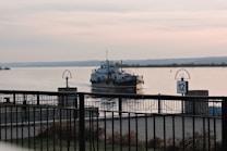 A boat is docked on a calm, wide river with a distant shoreline visible under a softly lit, cloudy sky. A metal fence runs along the foreground, and dock signs with the number 12 are displayed prominently.