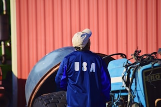 A person wearing a blue jacket with 'USA' printed on the back is standing in front of a tractor. The background includes a corrugated red metal wall and a large tire.