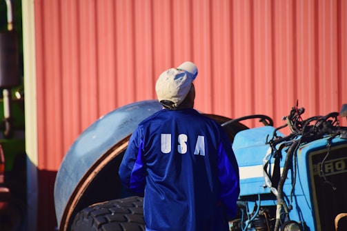 A person wearing a blue jacket with 'USA' printed on the back is standing in front of a tractor. The background includes a corrugated red metal wall and a large tire.