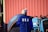 A person wearing a blue jacket with 'USA' printed on the back is standing in front of a tractor. The background includes a corrugated red metal wall and a large tire.