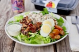 A plate of Cobb salad featuring fresh lettuce, sliced hard-boiled eggs, cherry tomatoes, diced turkey, crispy bacon, and crumbled blue cheese. A small container of creamy dressing is placed beside the salad on the plate. In the background, a can of La Croix and a takeout container can be seen.