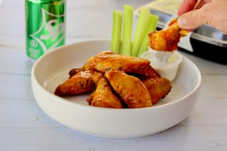 A white bowl contains several glazed chicken wings, with a hand dipping one wing into a container of creamy dipping sauce. Behind the bowl, there are sticks of celery standing upright. A green can of Sprite is placed nearby, partially visible. The surface is light-colored, providing a contrasting background for the food items.