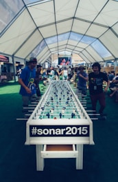 A large foosball table is set up under a spacious, airy tent with people engaged in playing or observing the game. The tent has a white, transparent ceiling, allowing natural light to filter through. In the background, a large screen displays an ongoing event, while individuals are casually dressed, creating a relaxed and lively atmosphere.