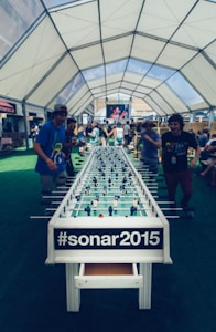 A large foosball table is set up under a spacious, airy tent with people engaged in playing or observing the game. The tent has a white, transparent ceiling, allowing natural light to filter through. In the background, a large screen displays an ongoing event, while individuals are casually dressed, creating a relaxed and lively atmosphere.